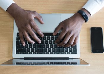 A black person working on his MacBook Pro while an iPhone placed on the wooden table